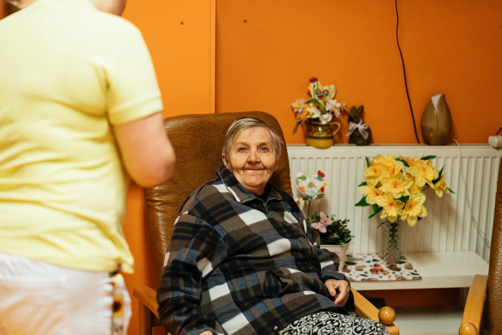 A senior woman in an armchair at home with her caretaker nearby — the scene of round-the-clock home care.